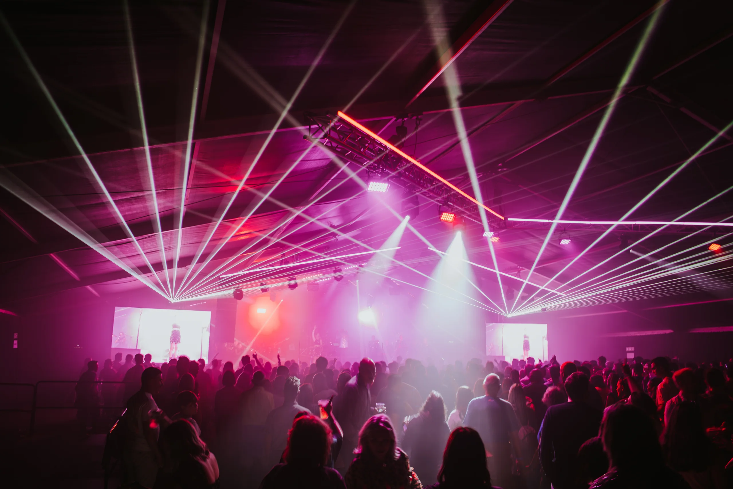 Crowd beneath an immersive display of magenta stage lighting and white laser beams